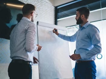 Consultant dressed in smart casual wear discussing ideas with a business team to improve systems and processes, in an office setting with a mock-up flip chart and whiteboard used for training
